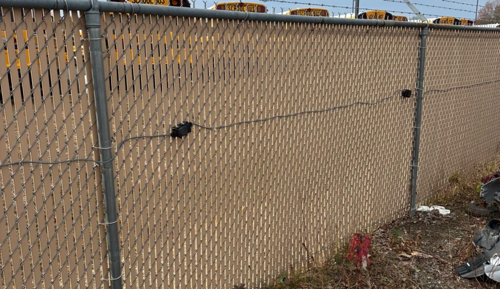 IRONCLAD sensor cable mounted with clips along a chain link fence with privacy slats and barbed wire at a vehicle impound lot.
