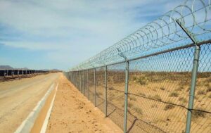 Chain link perimeter fence topped with concertina coil running into the distance across desert terrain, with solar panel rows and a service road alongside, at a solar farm in northern Mexico protected by RaySense fiber optic sensing.