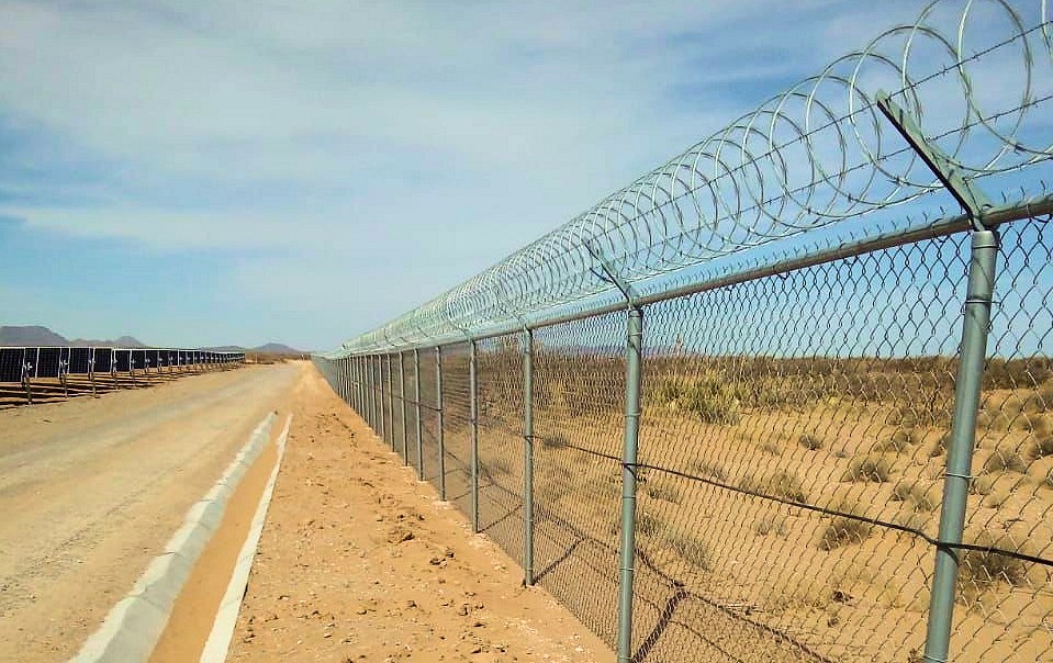 Chain link perimeter fence topped with concertina coil running into the distance across desert terrain, with solar panel rows and a service road alongside, at a solar farm in northern Mexico protected by RaySense fiber optic sensing.