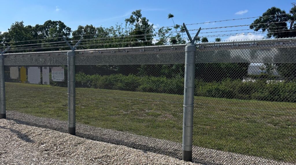 Chain link substation perimeter fence with barbed wire outriggers and an IRONCLAD sensor cable running along the fence fabric at a US electric utility substation.